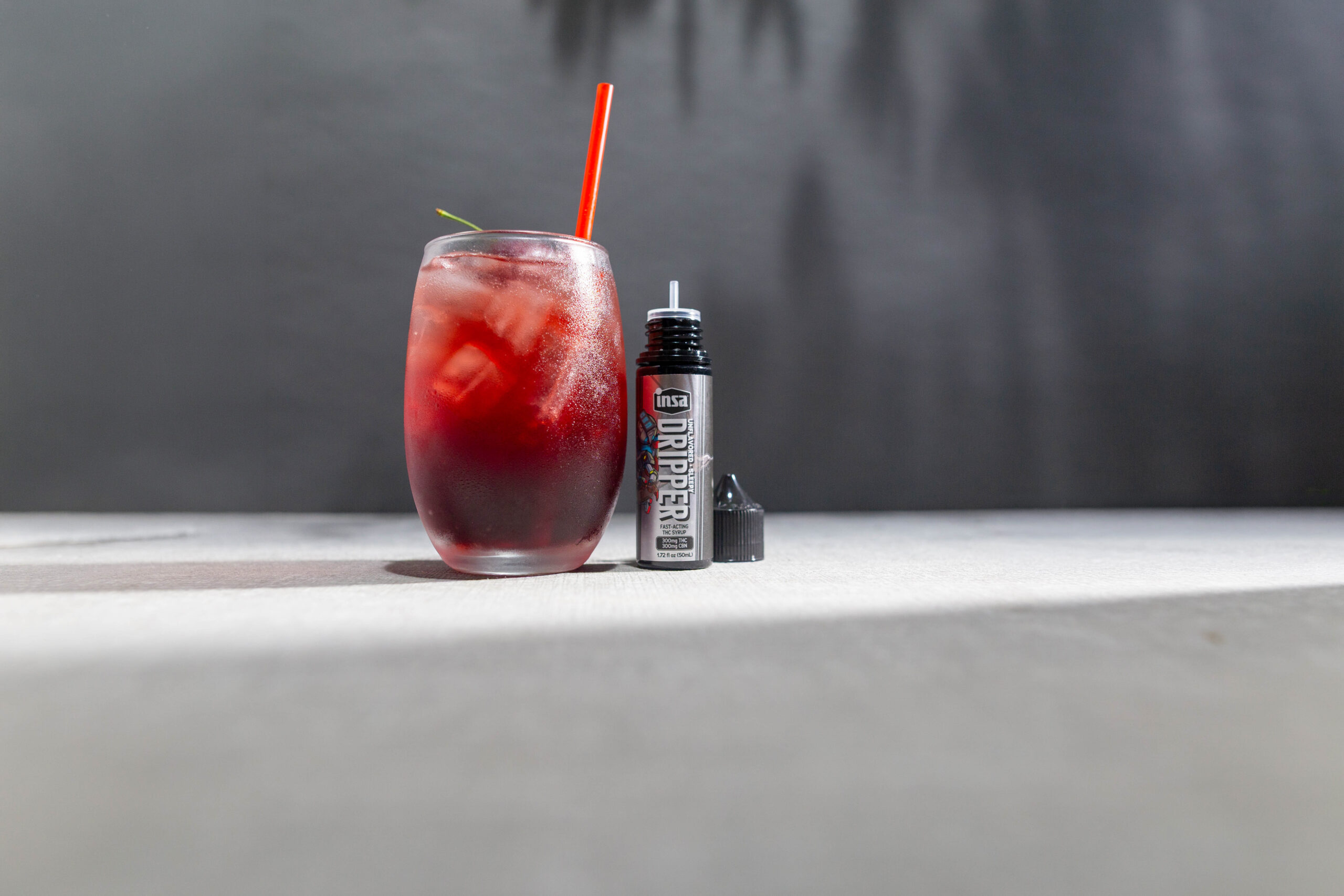 Cherry Moonlight Spritz: A red drink in a glass of ice with a cherry and straw, next to a bottle of Insa unflavored drippers with it's cap off and beside it