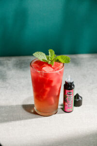 A Watermelon Wave Refresher in a tall glass with ice and mint leaf garnish, next to an uncapped bottle of Insa Watermelon Dripper