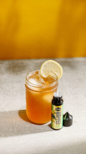 A Caddy Cooler drink in a glass with a lemon wedge next to a bottle of Insa Lemonade Drippers with the cap off